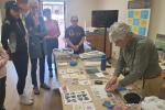 A woman demonstrates printmaking techniques to a small group gathered around a table filled with art prints and supplies in a bright room.