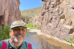 Hiker wearing a hat standing beside a narrow desert canyon stream, with tall rock walls rising on both sides under a bright blue sky.
