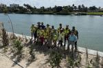 Group of people wearing safety vests standing on sandy shore near a body of water with greenery and buildings in the background.