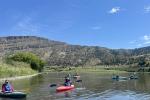 People in kayaks float on a river with hills in the background under a blue sky