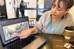 photo of a woman pointing to an illustrated dinosaur scene on a laptop computer screen