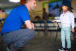 A person in a blue shirt crouches and talks with a small child wearing a backward cap indoors.