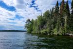 A scenic view of a forested shoreline under a partly cloudy sky, with calm lake water in the foreground.