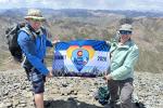 Two people stand on a rocky summit holding a flag reading "Summit 2026"
