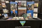 A person sits behind an informational booth for the Bureau of Land Management’s Eastern States office at a convention or expo. The table is covered with brochures, booklets, and educational materials. Large display banners behind the booth show outdoor scenes, wildlife, and public lands messaging. Two tall racks on either side hold additional publications. The person is seated at the center of the booth, smiling toward the camera.