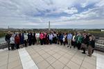 A large group of people stands together on a rooftop terrace with the Washington Monument and surrounding cityscape visible in the background under a partly cloudy sky.