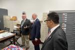 Three people stand together in a room with map drawers and archival furniture. One person speaks while holding papers, and the others listen attentively. Colorful documents and historical materials are laid out on nearby tables.