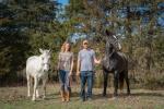  A photo showing (left to right) a white horse; a blonde woman with a t-shirt and jeans tucked into cowboy boots; a bearded man wearing a ballcap, t-shirt, and jeans; and a large dark horse with a white stripe down his face. They are standing in front of a wall of tall cedar trees.
