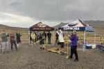 A couple dozen people stand around clapping while a person cuts a yellow ribbon; a cloudy desert landscape behind