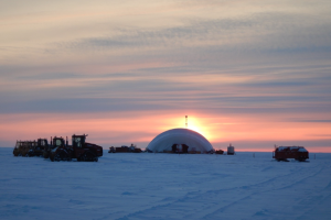 Snow-covered ground, with an orange sunset behind a large inflatable dome. Heavy equipment nearby.