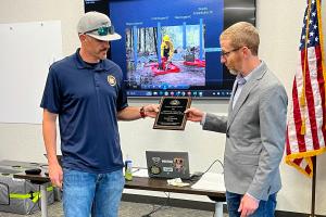 Austin Blackett, left, is handed a plague from Matt Preston, right. Austin is a slimmer, middle aged male with a dark mustache wearing blue jeans. Matt is a slim, middle aged male standing slightly shorter than Austin. Matt is wearing a gray suit jacket with blue jeans.