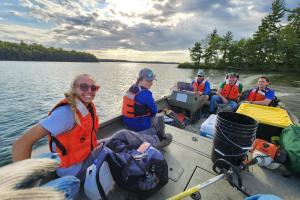 A group of individuals wearing orange life jackets sits in a boat on a lake, surrounded by trees and a cloudy sky.