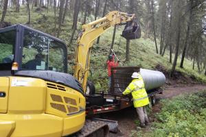 Workers using a yellow excavator to unload a large metal culvert from a trailer on a forest road.