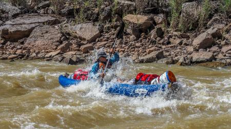 A man in a blue inflatable kayak paddles through rough rapids with white water spraying all around him.