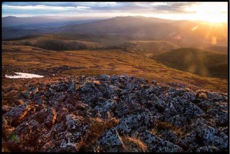 Alpine rocks above the ridges and valleys of the Fortymile Wild and Scenic River.  The sun is low over the mountains creating a flare coming out of the upper right of the image. 