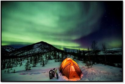 Winter scene in the White Mountains National Recreation Area with the green glow of the aurora in the sky. A small yellow tent is lit up. Snow shoes are placed near the tent. Small spruce trees line the rounded, snow ridge line.
