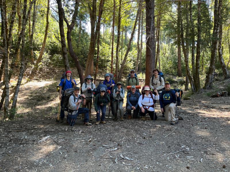 group of people stand in a wooded area near a trail head sign.