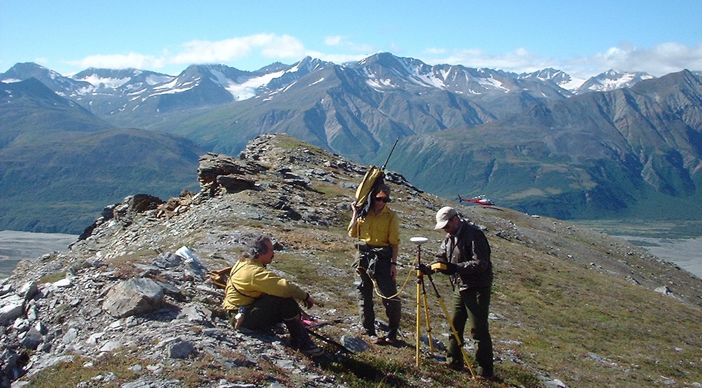 Three cadastral surveyors work on the top of a mountain with a landscape view. BLM photo.
